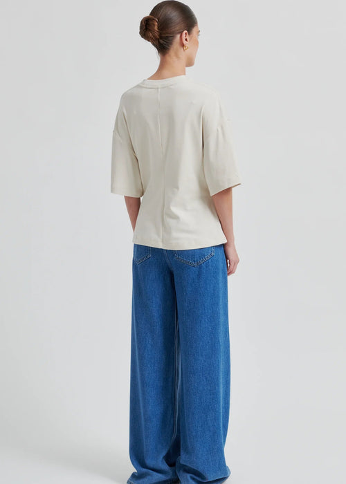 Woman wearing a cream-colored top with a brooch and blue jeans on a white background