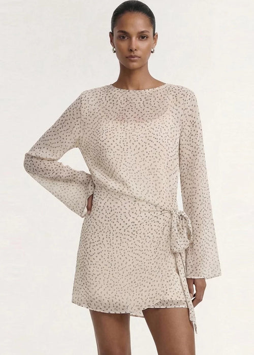 A model wearing a long-sleeve, wrap-style mini dress with a delicate beige and white floral print, set against a plain off-white studio background.