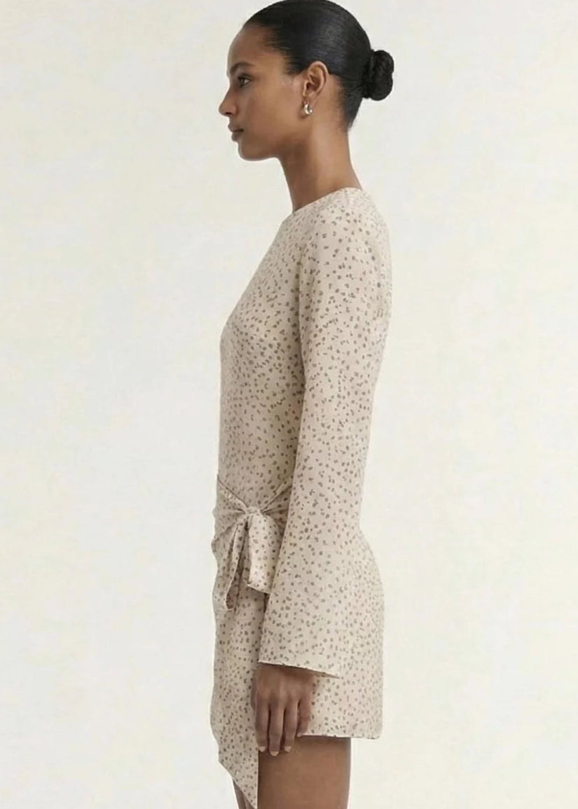 A model wearing a long-sleeve, wrap-style mini dress with a delicate beige and white floral print, set against a plain off-white studio background.