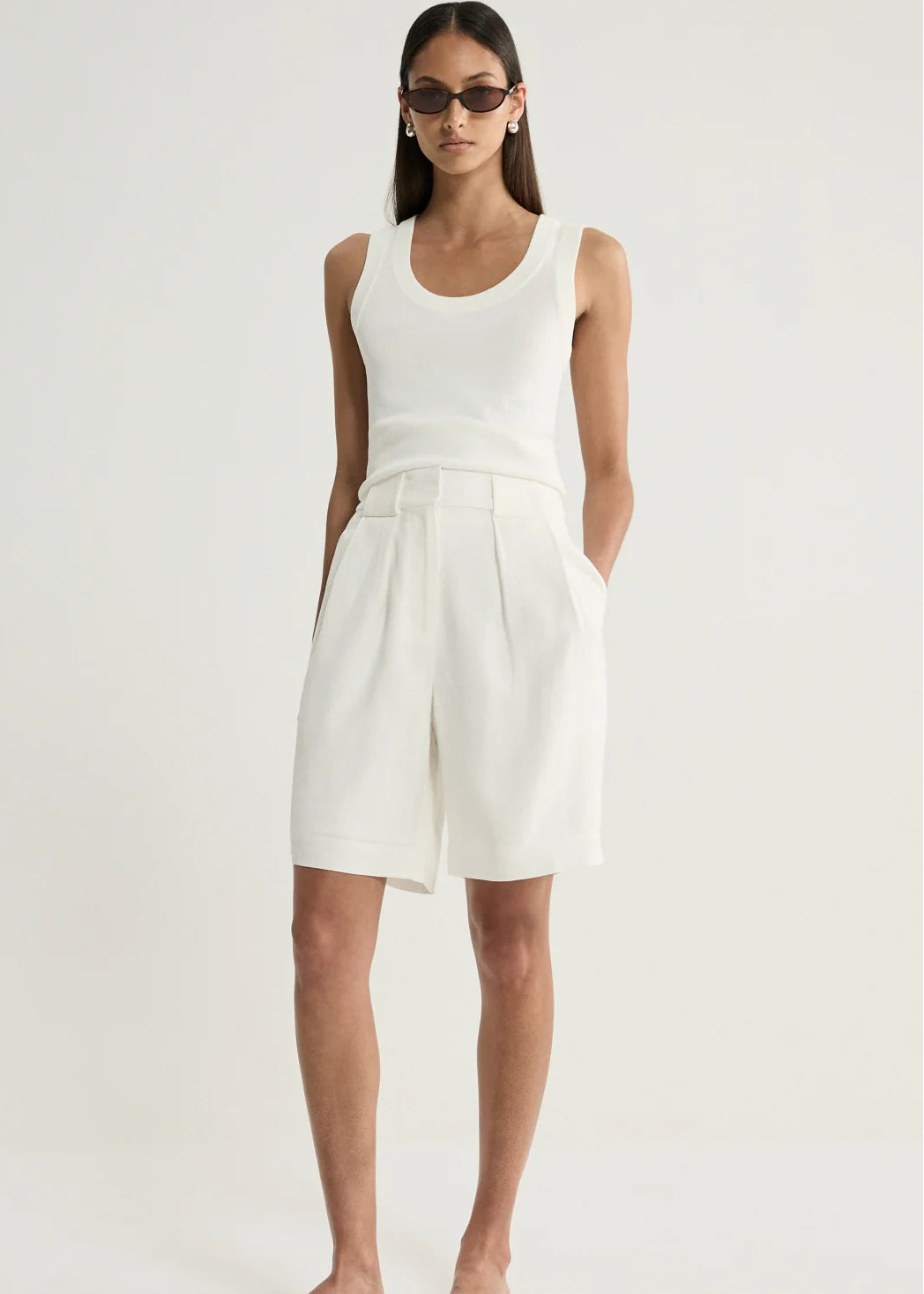 A model wearing a white sleeveless knit top and matching white pleated shorts, styled with black sunglasses and black slide sandals against a plain white studio background.