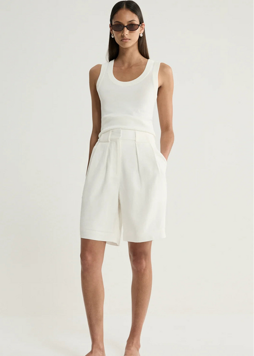 A model wearing a white sleeveless knit top and matching white pleated shorts, styled with black sunglasses and black slide sandals against a plain white studio background.