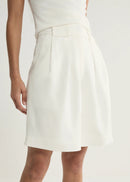 A model wearing a white high-waisted pleated skirt with a matching belt, paired with a white top, shown from the waist down against a light off-white background.
