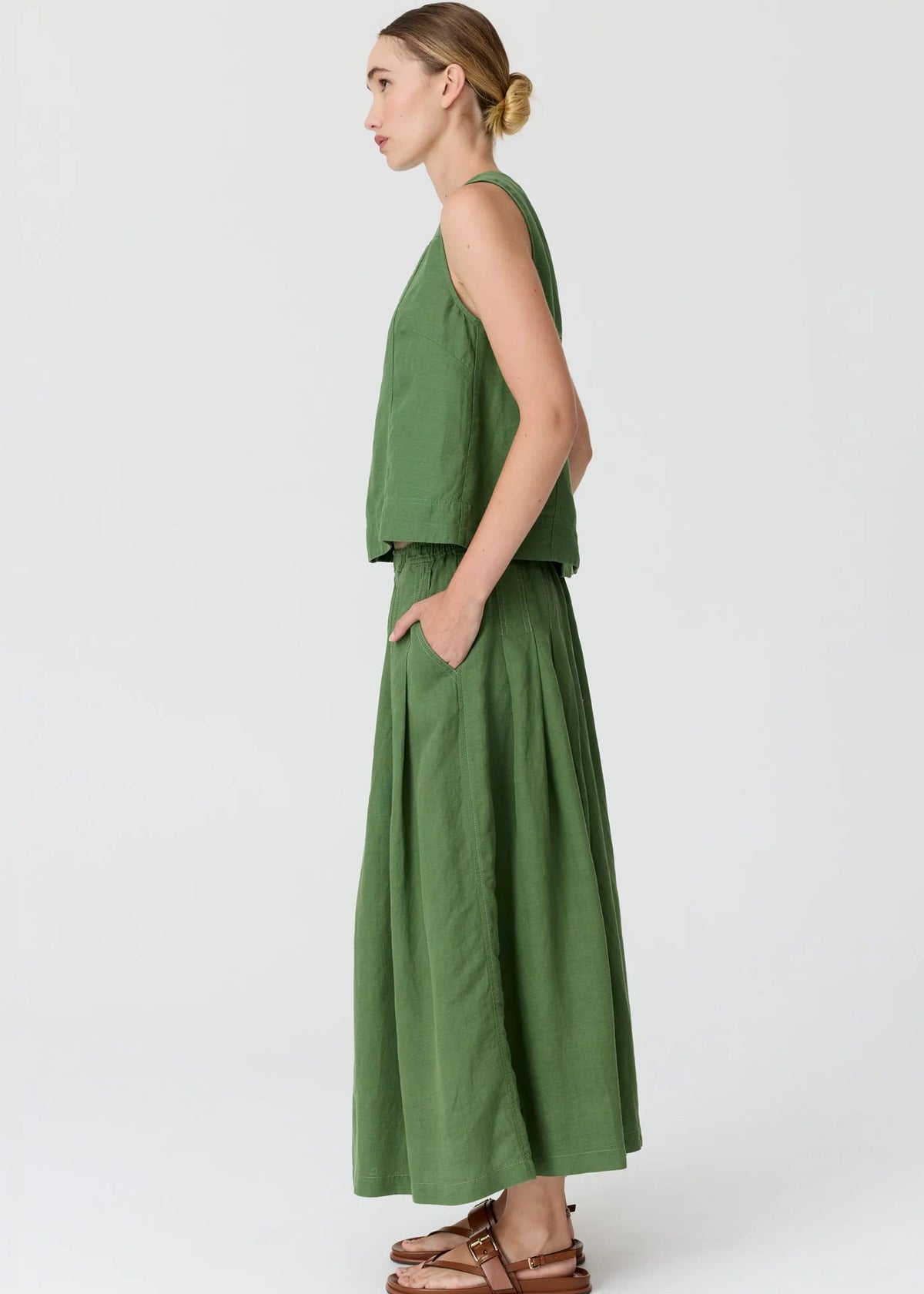 A woman modelling in profile a matching olive green two-piece linen set consisting of a sleeveless crop top and a high-waisted, wide-leg maxi skirt, paired with brown leather sandals against a light grey studio background.