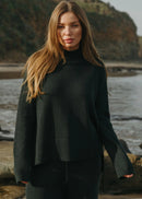 Woman wearing a dark green sweater standing by a body of water with a natural landscape.
