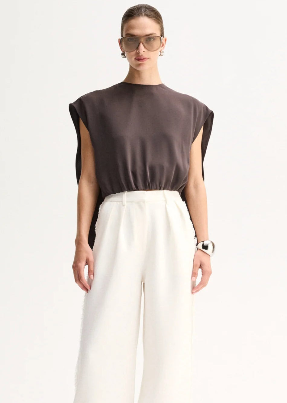 A model stands against a plain white studio background wearing a dark brown sleeveless top with wide legs and high-waisted white wide-leg trousers.