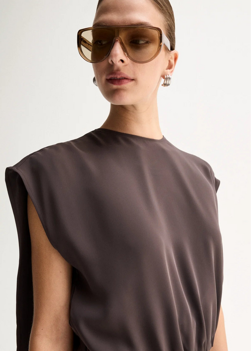 A close-up of a model wearing a dark brown sleeveless top and oversized brown-tinted sunglasses against a light grey background.