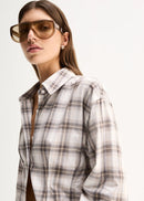 A woman modelling a long-sleeve plaid button-up shirt in beige, white, and grey, paired with white wide-leg pants and black sandals, against a plain white studio background.