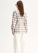 A woman viewed from the back wearing a cream and brown plaid button-up shirt, white wide-leg pants, and black pointed flats against a plain white studio background.
