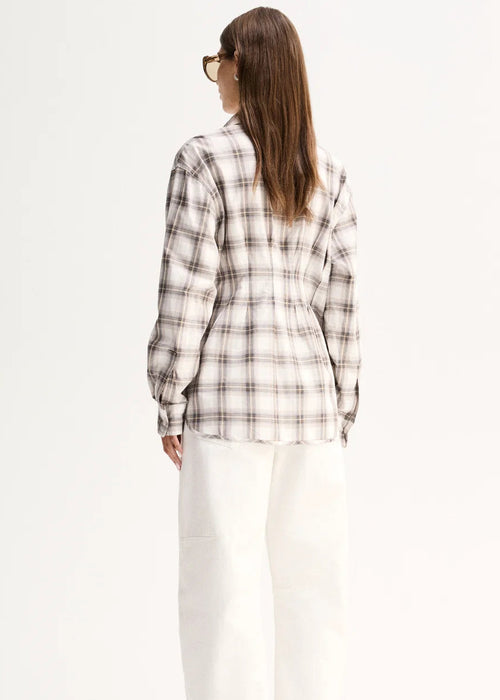 A woman viewed from the back wearing a cream and brown plaid button-up shirt, white wide-leg pants, and black pointed flats against a plain white studio background.