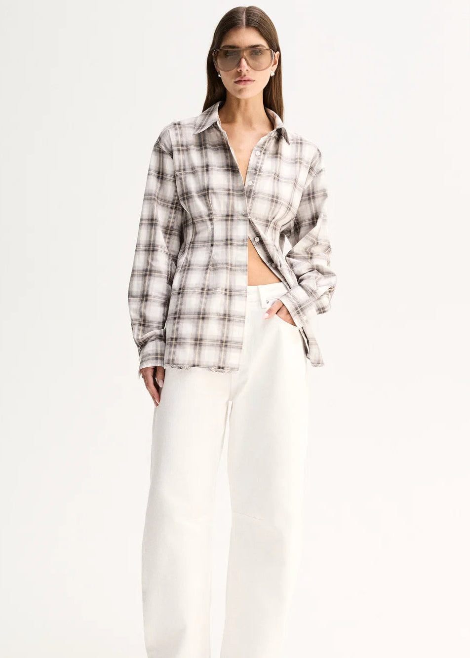 A woman modelling a long-sleeve plaid button-up shirt in beige, white, and grey, paired with white wide-leg pants and black sandals, against a plain white studio background.