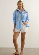 A woman modelling a light blue button-up shirt with white floral embroidery on the shoulders, paired with white shorts and brown sandals, standing against a neutral studio background.
