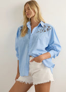 A woman modelling a light blue button-up shirt with white floral embroidery on the shoulders, paired with white shorts and brown sandals, standing against a neutral studio background.