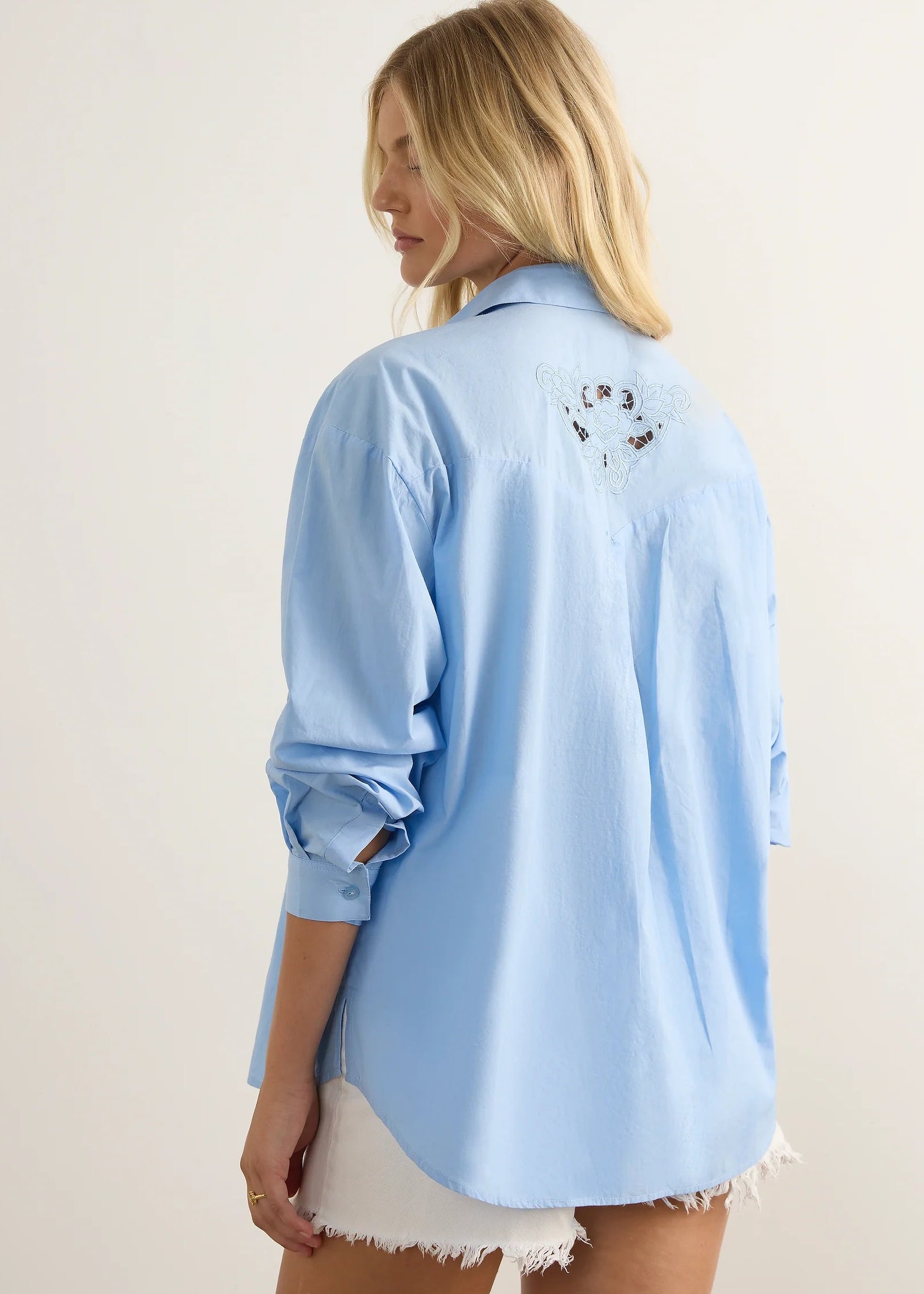 A woman modelling a light blue button-up shirt with white floral embroidery on the shoulders, paired with white shorts and brown sandals, standing against a neutral studio background.