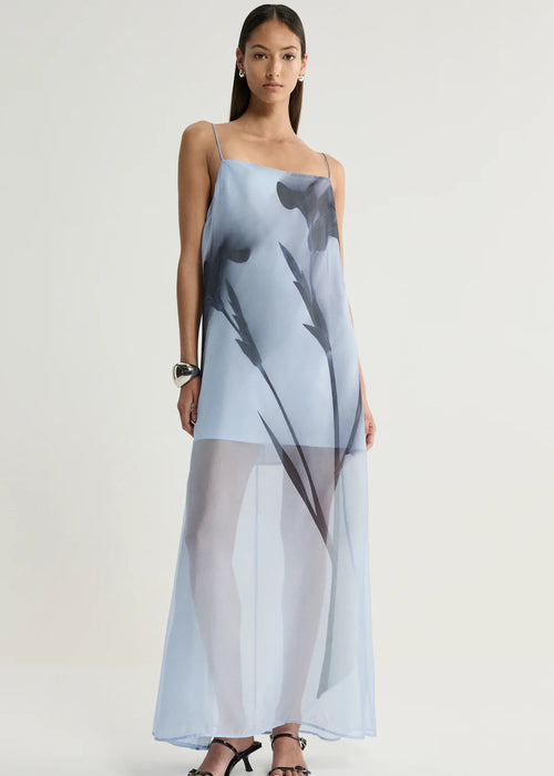 A woman modelling a sheer, light blue maxi dress with a large black abstract print, photographed against a neutral light grey studio background.