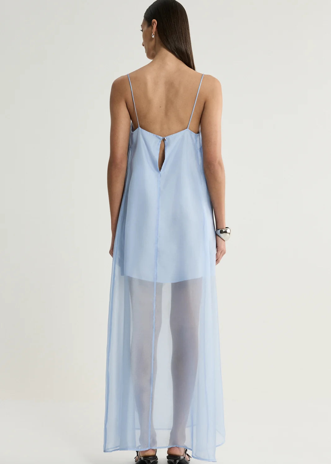 A woman shown from the back wearing a light blue sheer maxi slip dress with thin straps and a keyhole back detail, paired with white tights and black heels, against a plain light grey studio background.