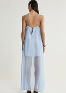 A woman shown from the back wearing a light blue sheer maxi slip dress with thin straps and a keyhole back detail, paired with white tights and black heels, against a plain light grey studio background.