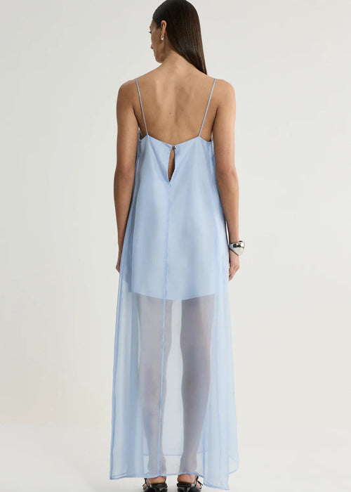 A woman shown from the back wearing a light blue sheer maxi slip dress with thin straps and a keyhole back detail, paired with white tights and black heels, against a plain light grey studio background.