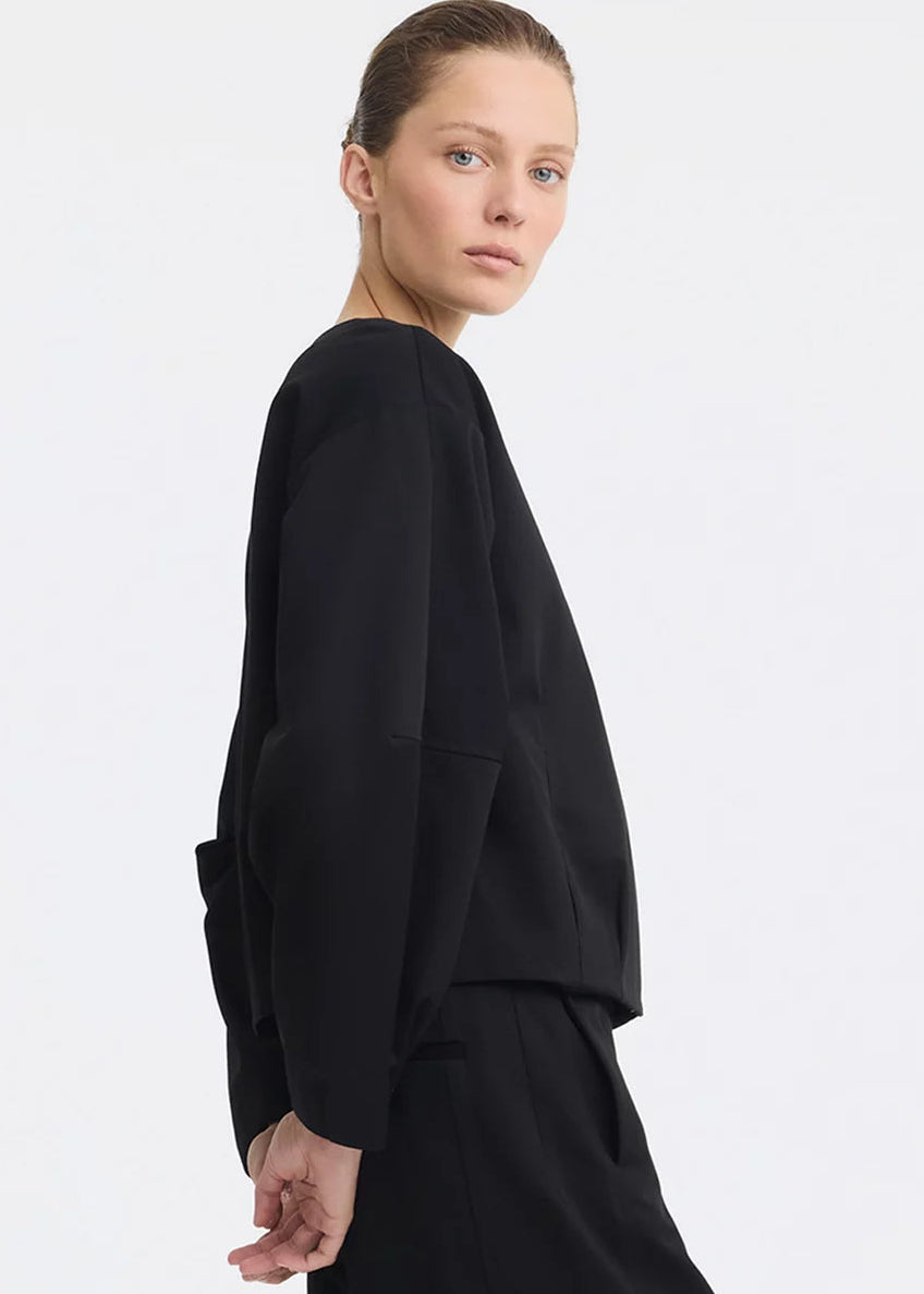 A model wearing a black cropped jacket with a front zipper and wide sleeves, paired with a black skirt, against a light grey studio background.