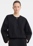 A model wearing a black cropped jacket with a front zipper and wide sleeves, paired with a black skirt, against a light grey studio background.