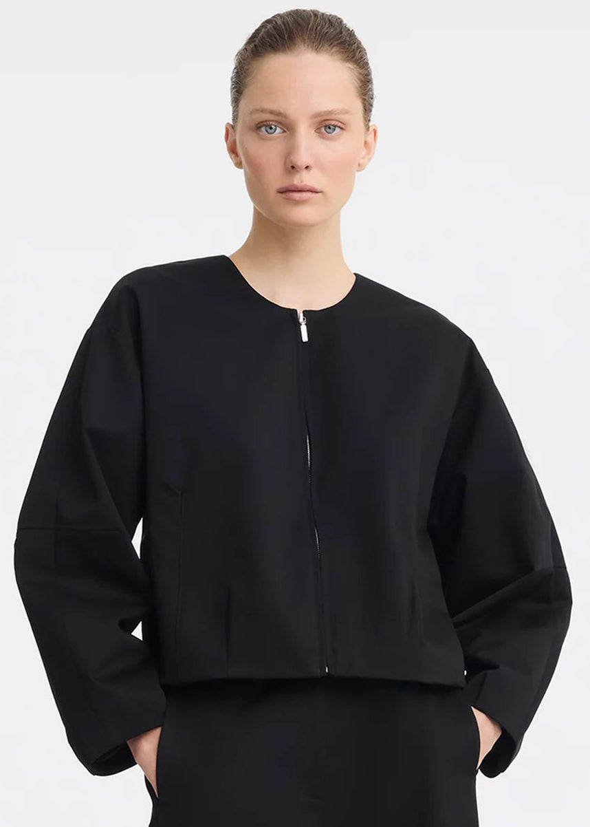 A model wearing a black cropped jacket with a front zipper and wide sleeves, paired with a black skirt, against a light grey studio background.