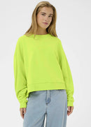 Woman wearing a bright green sweater and light blue jeans against a white  wall.