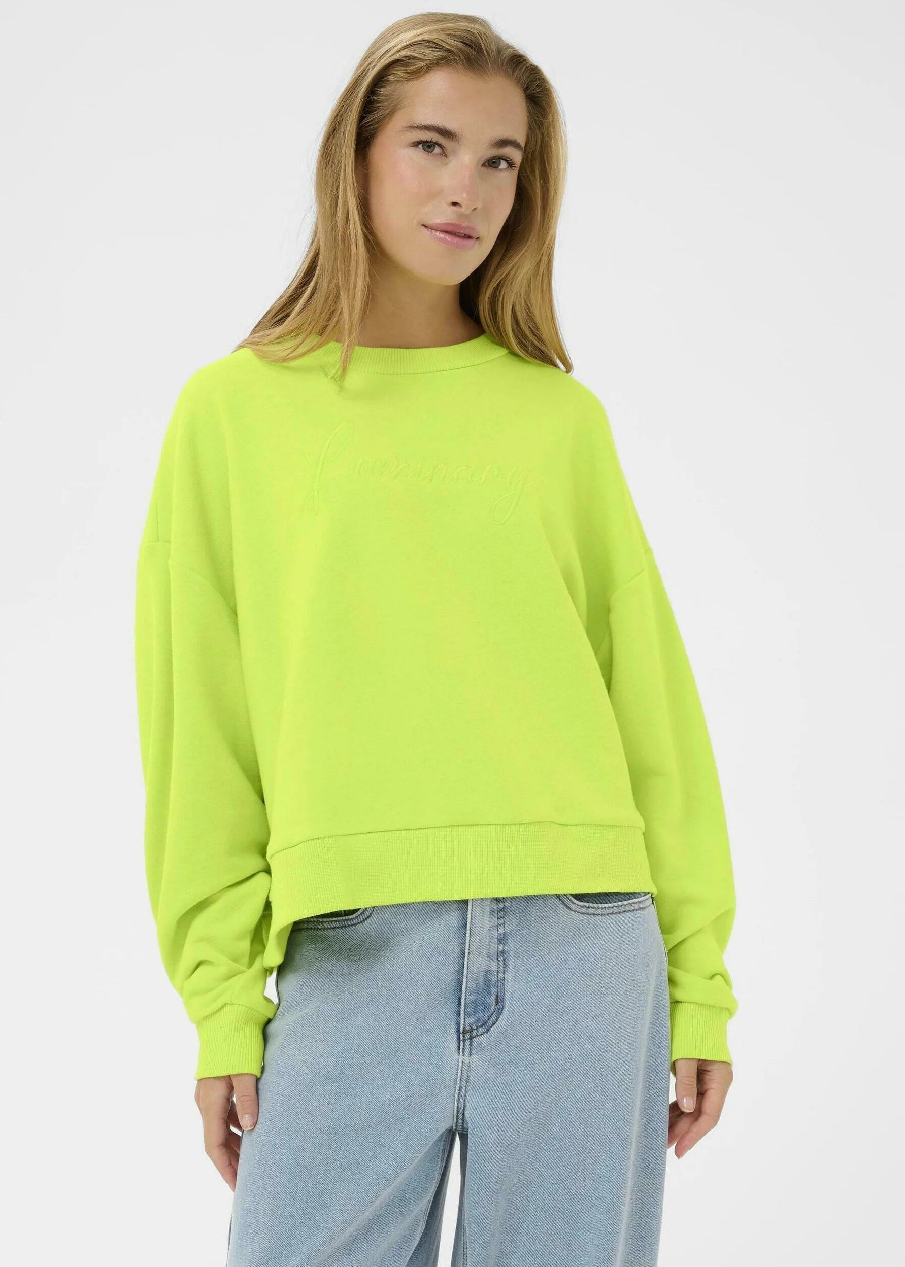 Woman wearing a bright green sweater and light blue jeans against a white  wall.