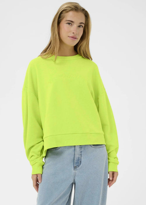 Woman wearing a bright green sweater and light blue jeans against a white  wall.