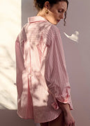 A woman wearing a pink and white vertically striped button-down shirt, photographed from the back in warm, directional sunlight against a neutral studio background.