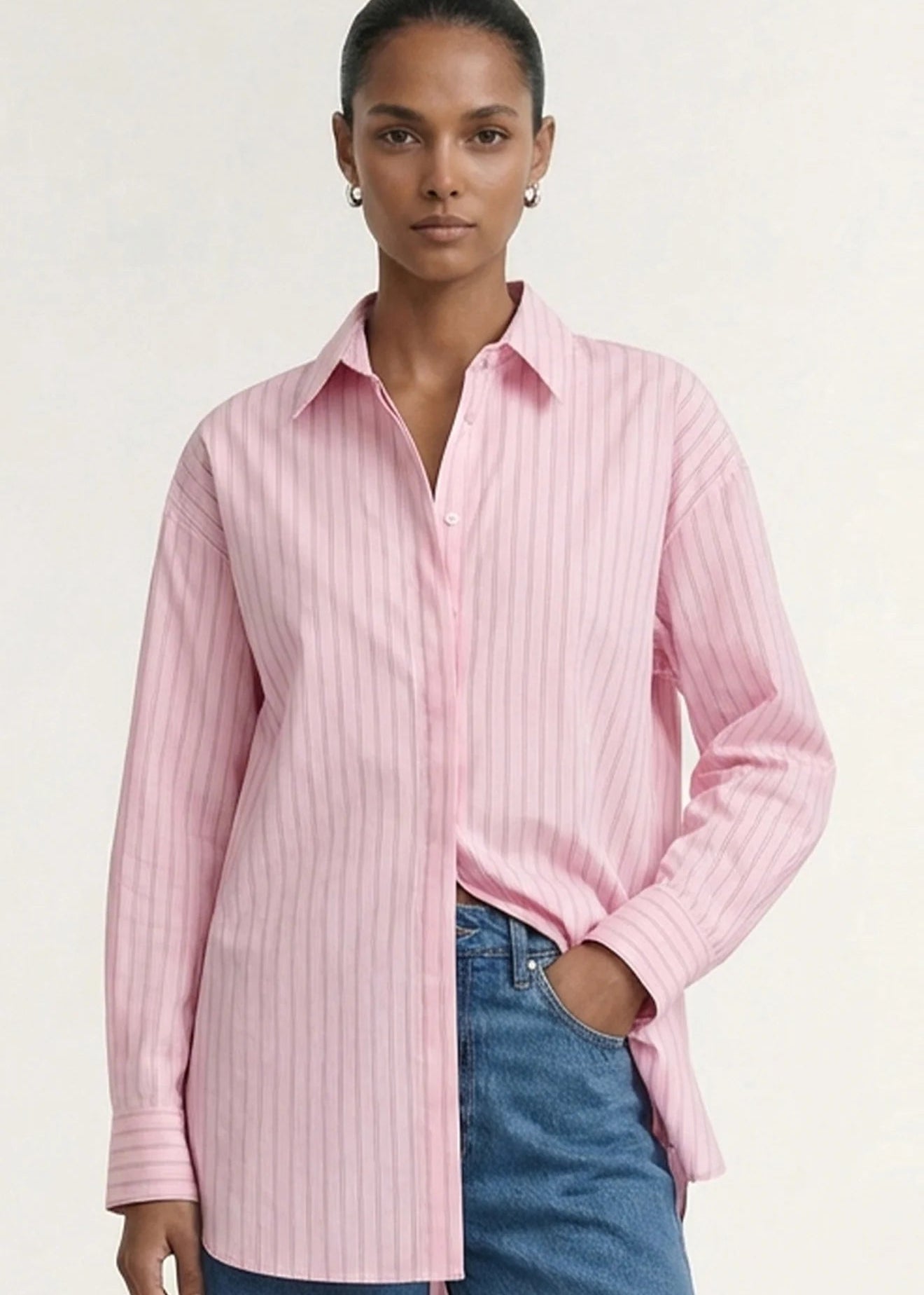 A woman modeling a pink and white vertically striped button-down shirt with blue jeans, photographed against a clean off-white studio background.