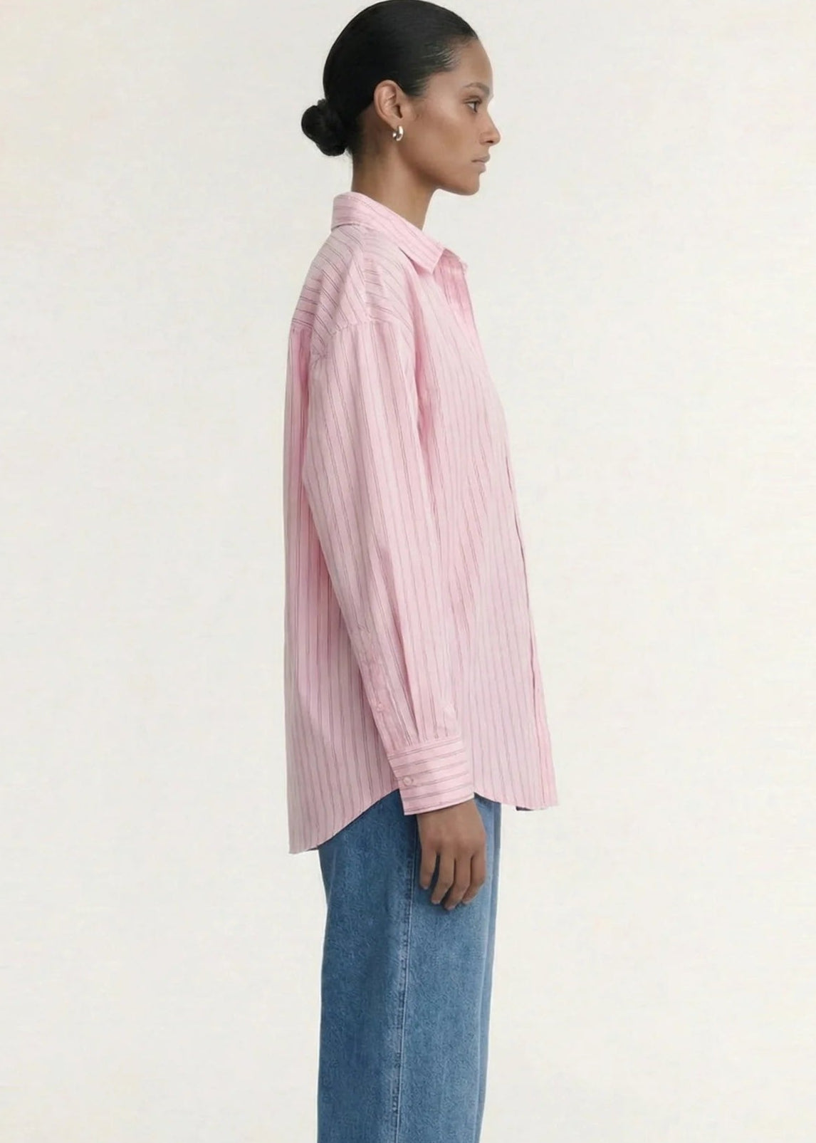 A woman modelling a pink and white vertically striped button-down shirt with blue jeans, photographed against a clean off-white studio background.