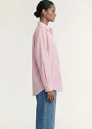 A woman modelling a pink and white vertically striped button-down shirt with blue jeans, photographed against a clean off-white studio background.