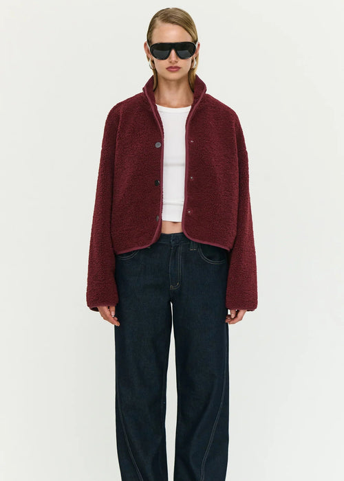 Woman wearing a burgundy jacket, white top, and dark jeans on a white background