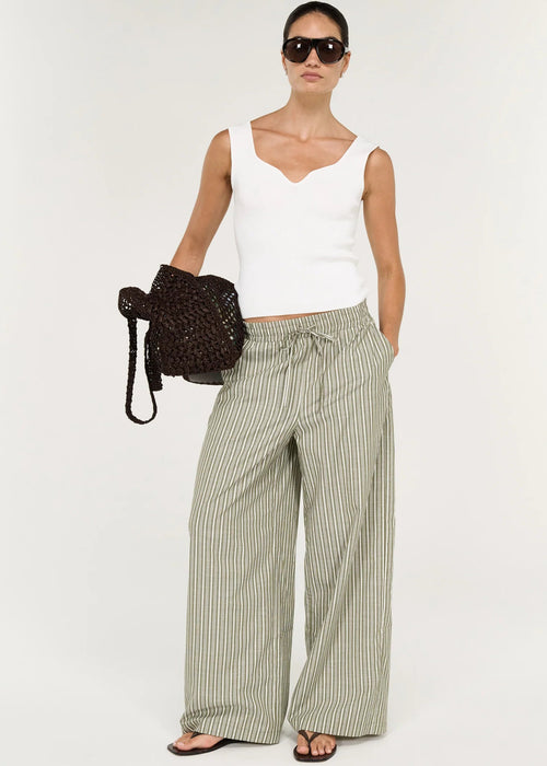 A model wearing a white sleeveless top, wide-leg striped trousers in beige and green, sunglasses, and holding a dark woven handbag against a plain white studio background.