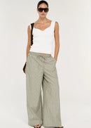 A model wearing a white sleeveless top, wide-leg striped trousers in beige and green, sunglasses, and holding a dark woven handbag against a plain white studio background.