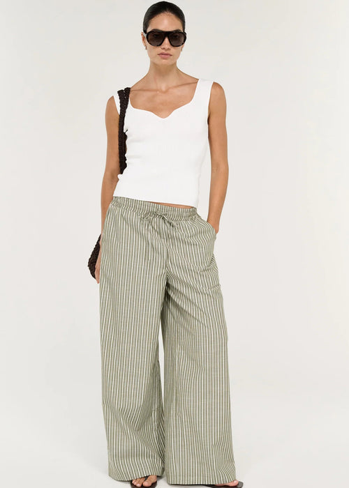 A model wearing a white sleeveless top, wide-leg striped trousers in beige and green, sunglasses, and holding a dark woven handbag against a plain white studio background.