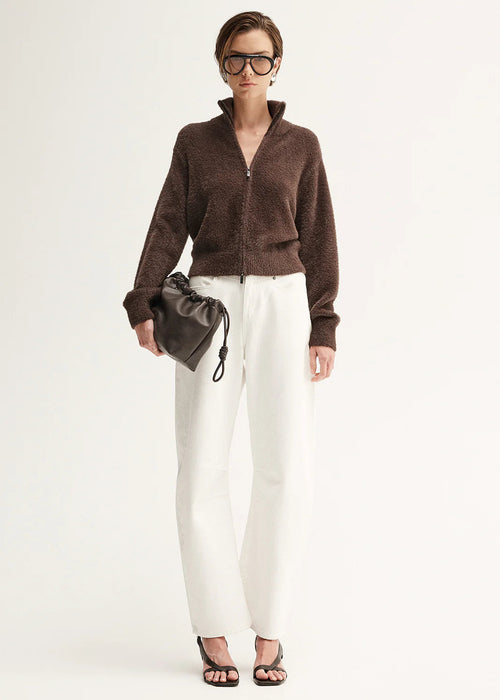 Woman wearing a brown cardigan, white pants, and holding a grey clutch against a white background