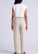 A woman modelling a white sleeveless top and beige wide-leg linen pants, holding a woven straw handbag, against a light grey studio background.