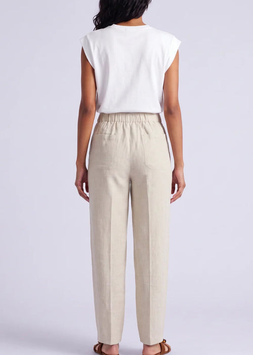 A woman modelling a white sleeveless top and beige wide-leg linen pants, holding a woven straw handbag, against a light grey studio background.