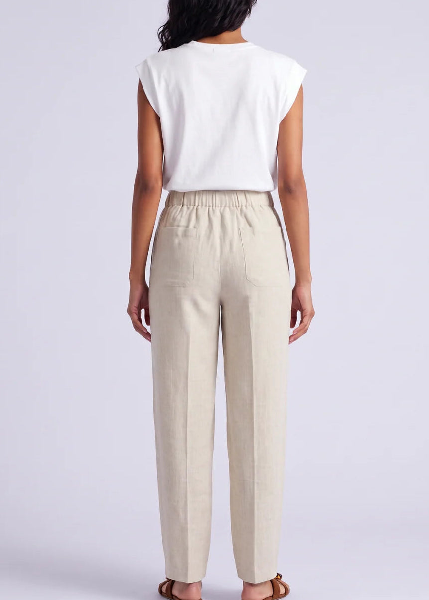A woman modelling a white sleeveless top and beige wide-leg linen pants, holding a woven straw handbag, against a light grey studio background.
