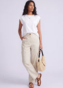 A woman modelling a white sleeveless top and beige wide-leg linen pants, holding a woven straw handbag, against a light grey studio background.