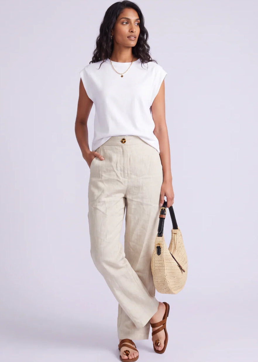 A woman modelling a white sleeveless top and beige wide-leg linen pants, holding a woven straw handbag, against a light grey studio background.