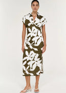 A woman modelling a short-sleeve midi dress with a bold white floral print on an olive green base, paired with white heeled thong sandals against a plain white studio background.