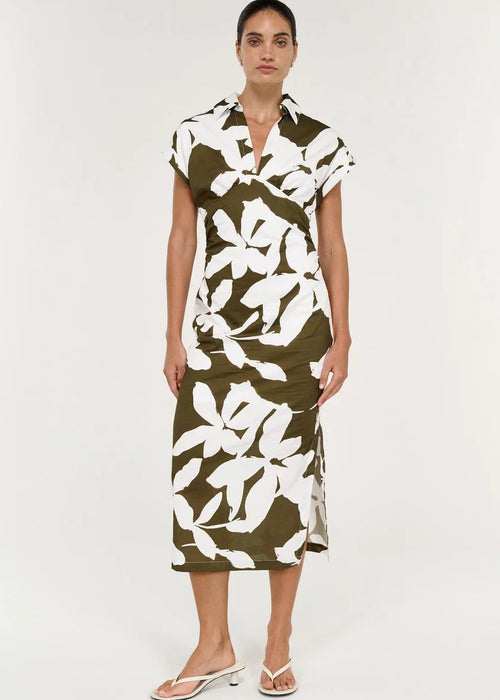 A woman modelling a short-sleeve midi dress with a bold white floral print on an olive green base, paired with white heeled thong sandals against a plain white studio background.