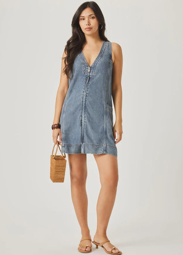 Woman wearing a denim dress holding a woven bag on a plain background