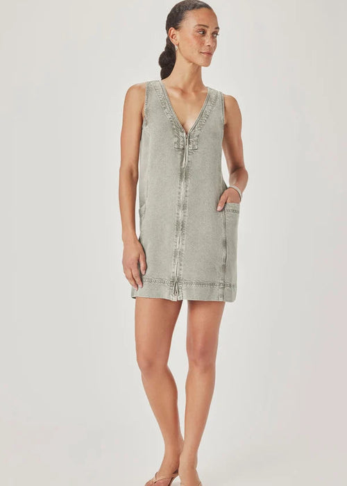 A woman modelling a sleeveless, V-neck mini dress with a faded green wash and decorative trim, paired with pink sandals, against a light grey studio background.