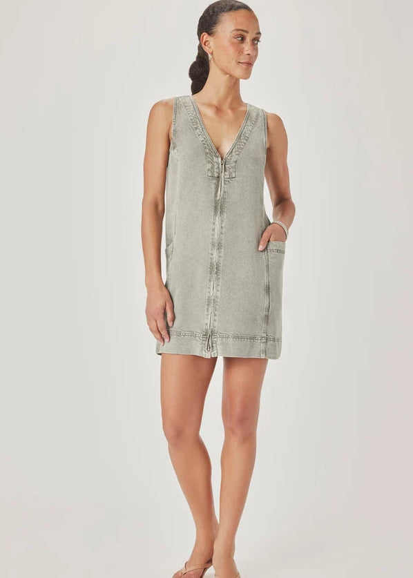 A woman modelling a sleeveless, V-neck mini dress with a faded green wash and decorative trim, paired with pink sandals, against a light grey studio background.