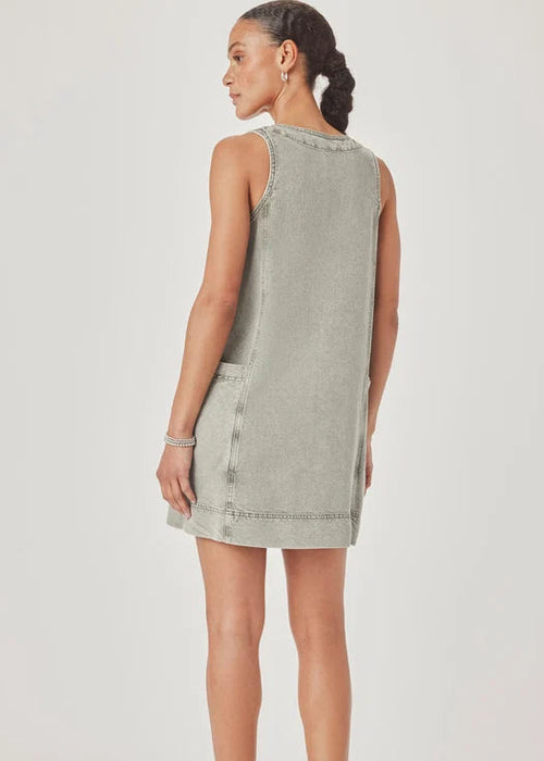 A woman modelling a sleeveless, V-neck mini dress with a faded green wash and decorative trim, paired with pink sandals, against a light grey studio background.