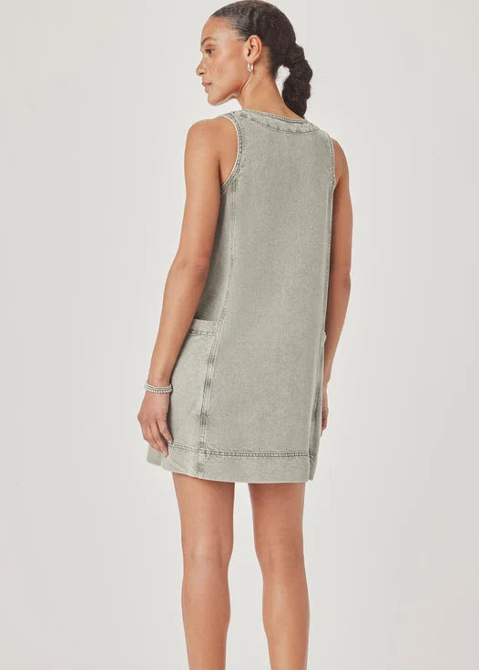 A woman modelling a sleeveless, V-neck mini dress with a faded green wash and decorative trim, paired with pink sandals, against a light grey studio background.