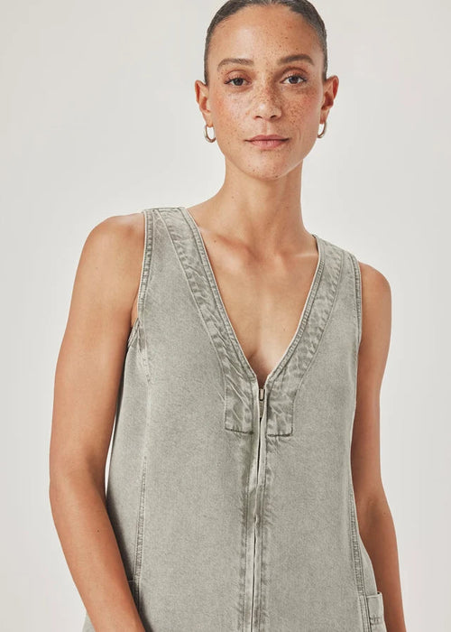A woman modelling a sleeveless, V-neck mini dress with a faded green wash and decorative trim, paired with pink sandals, against a light grey studio background.