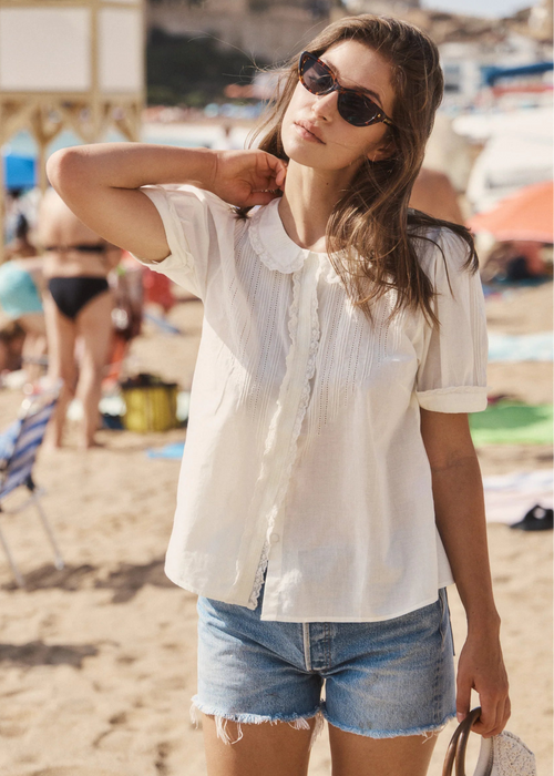 A woman stands on a sandy beach wearing a white short-sleeve button-front blouse with a ruffled collar, light blue denim cut off shorts, and dark sunglasses.
